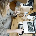Empreendedora fotografa sapatos com o celular em um espaço de trabalho, ao lado de um laptop e de pacotes para envio.