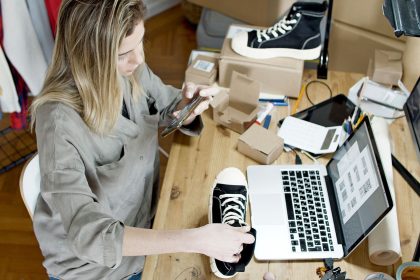 Empreendedora fotografa sapatos com o celular em um espaço de trabalho, ao lado de um laptop e de pacotes para envio.