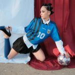 Modelo posa ao ar livre com uma bola de futebol e sapatos de salto, em uma composição que mistura esporte e moda.