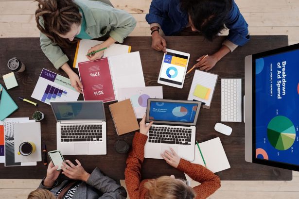 Equipe diversa de marketing digital colabora em uma mesa usando laptops e tablets.