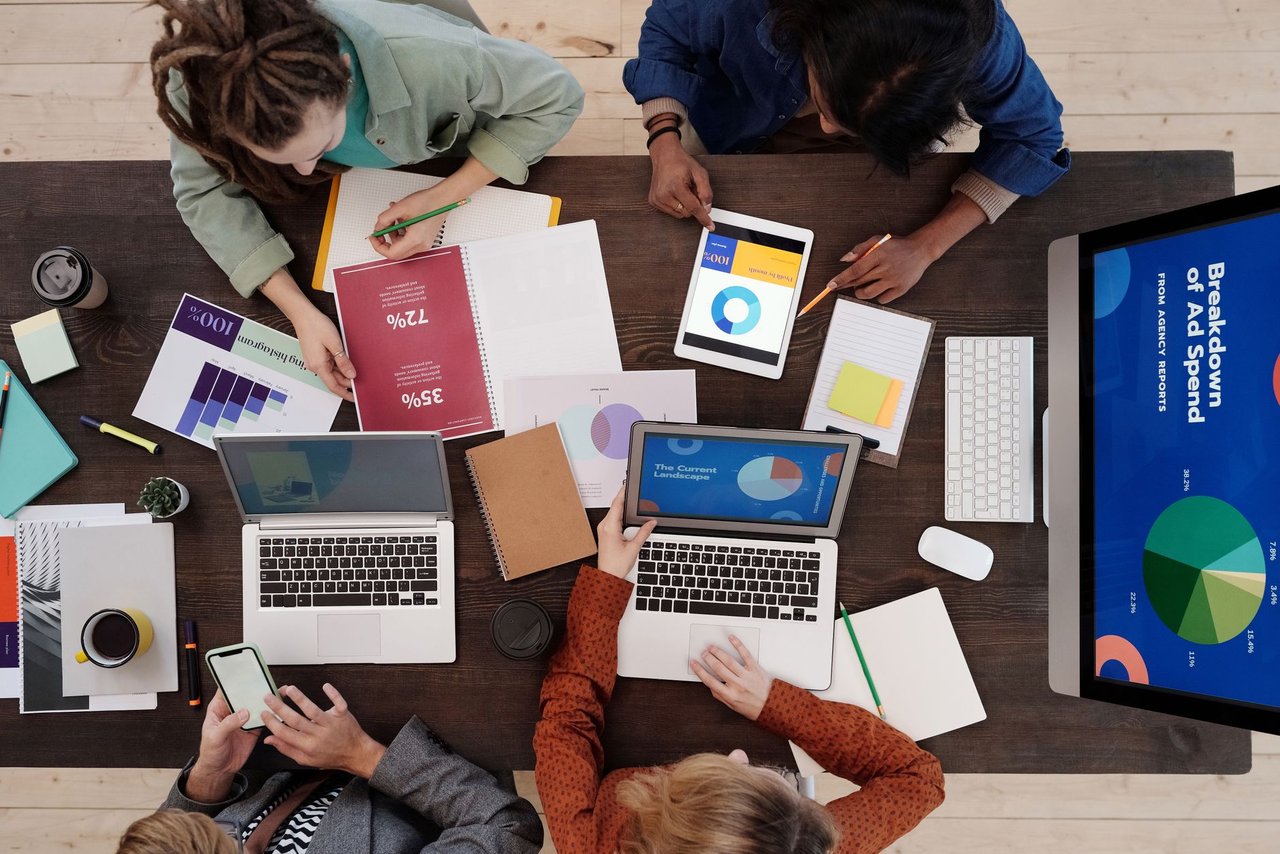 Equipe diversa de marketing digital colabora em uma mesa usando laptops e tablets.