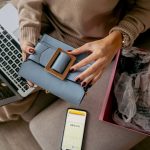 Mulher usa um laptop enquanto desembrulha uma bolsa comprada pela internet em casa.