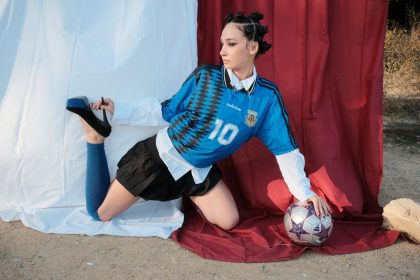 Modelo posa ao ar livre com uma bola de futebol e sapatos de salto, em uma composição que mistura esporte e moda.