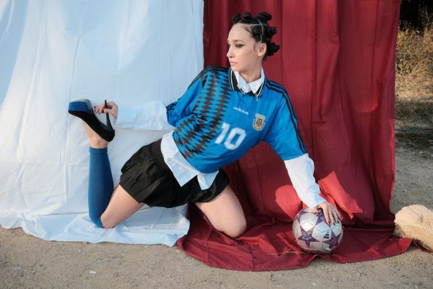 Modelo posa ao ar livre com uma bola de futebol e sapatos de salto, em uma composição que mistura esporte e moda.