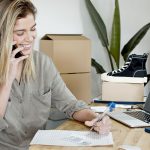 Empreendedora usa o celular e o notebook enquanto organiza anotações e pedidos de sua loja virtual.