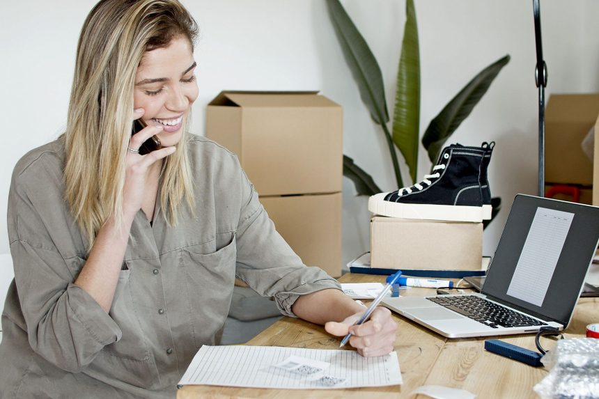 Empreendedora usa o celular e o notebook enquanto organiza anotações e pedidos de sua loja virtual.